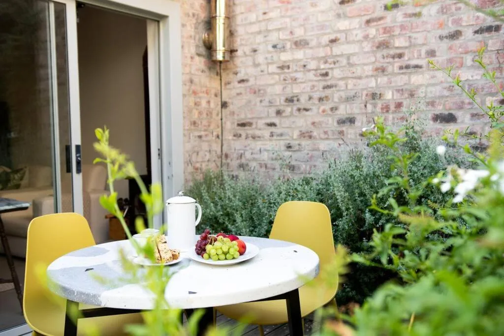 Outdoor patio with round white table, yellow chairs, fresh fruit, and potted plants
