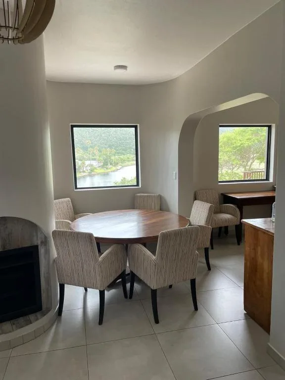 Modern dining area with round wooden table and upholstered chairs, large windows overlooking lagoon