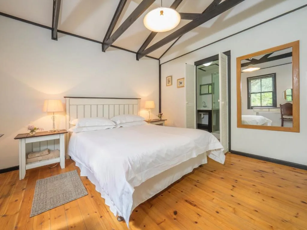 Bright bedroom with white bed, wooden beams, and ensuite bathroom visible