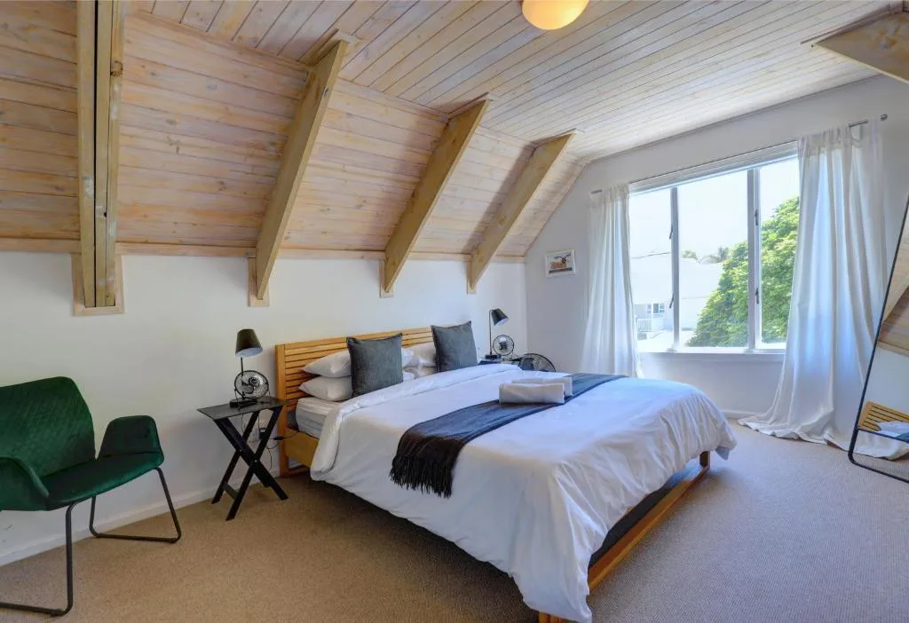 Bright attic bedroom with wooden beams, queen bed, green chair, and windows