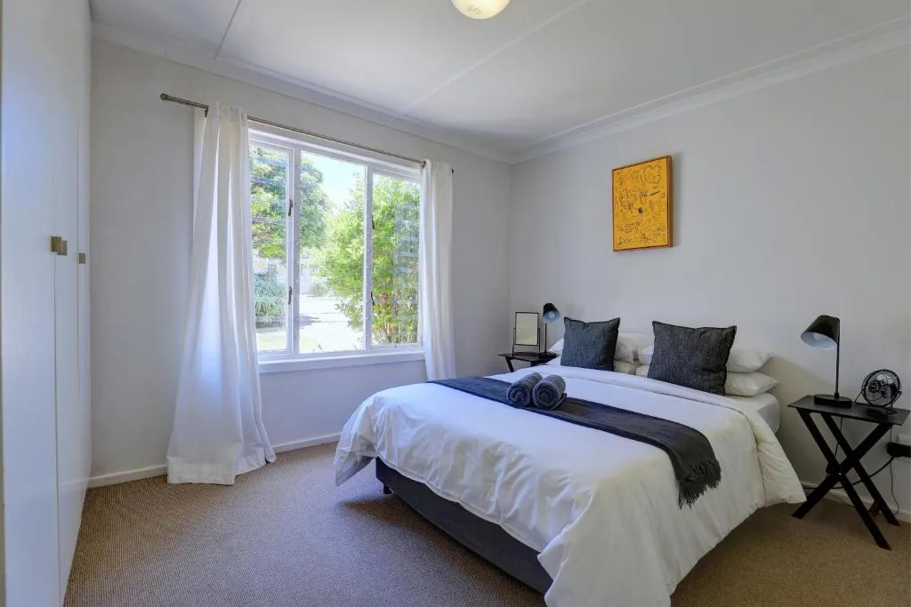 Bright bedroom with queen bed, white bedding, dark pillows, and garden views through windows