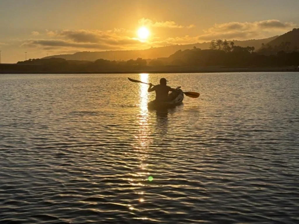 Kayaker paddling on golden sunset lagoon with forested hills beyond