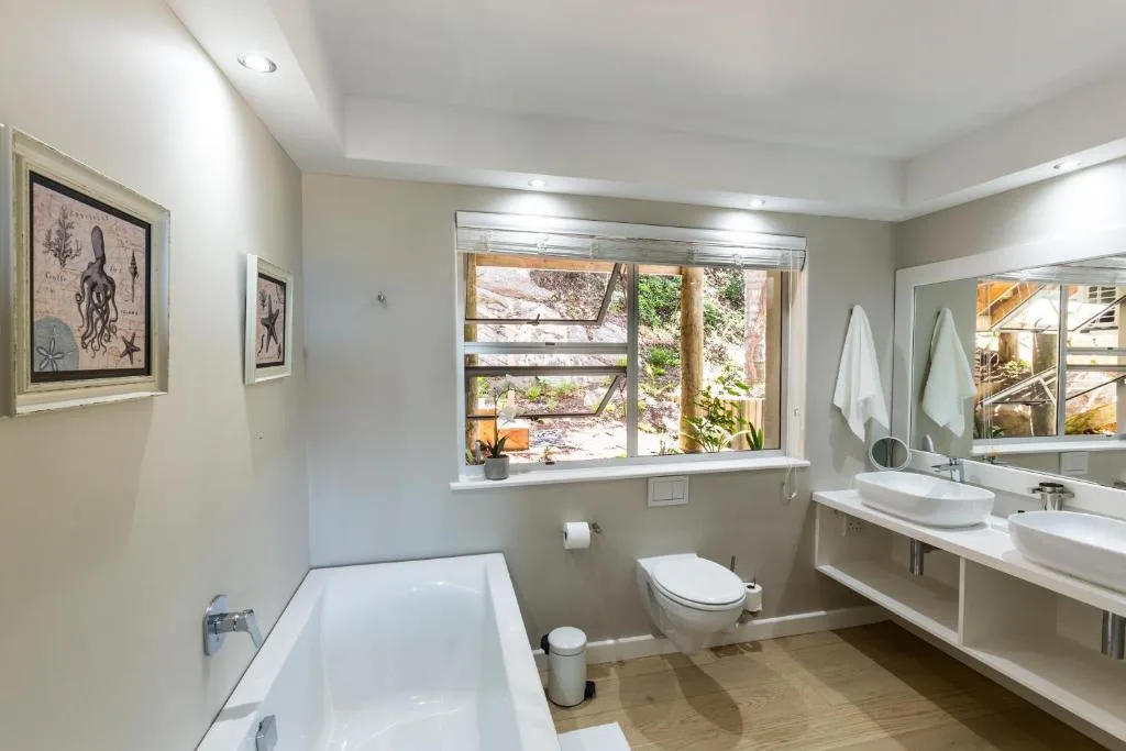Spacious bathroom with bathtub, dual vanity sinks, toilet, and garden views
