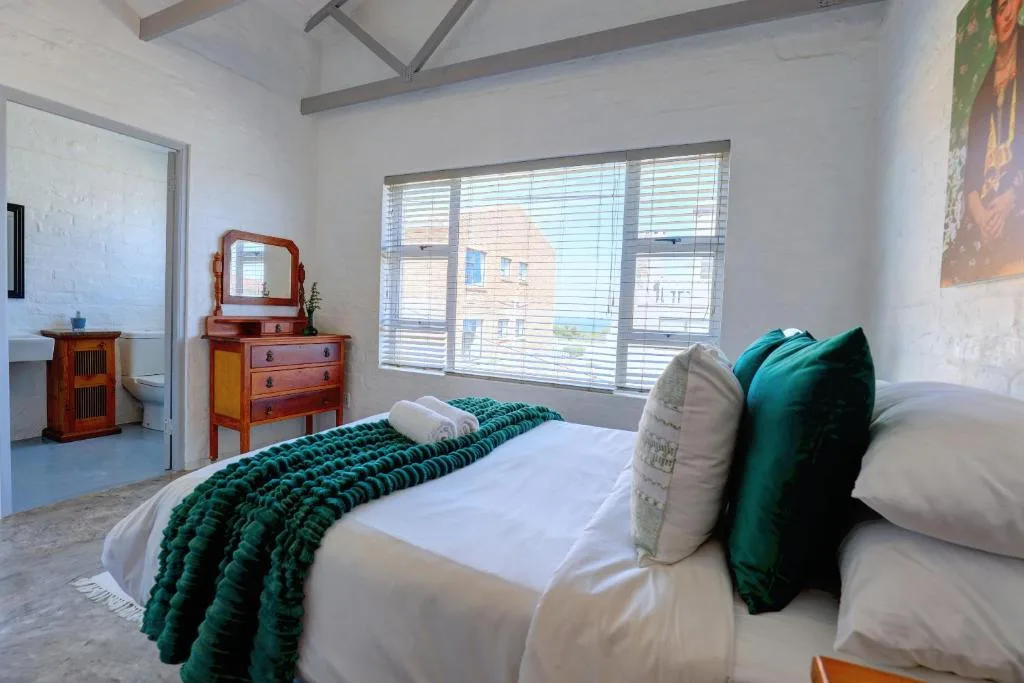 Bright bedroom with double bed, green accents, wooden dresser, and large windows