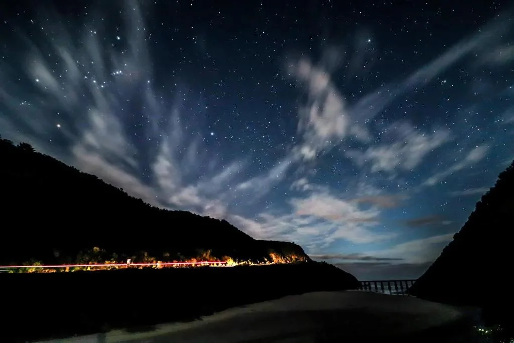 Starlit night sky above illuminated coastal village nestled between dark mountains