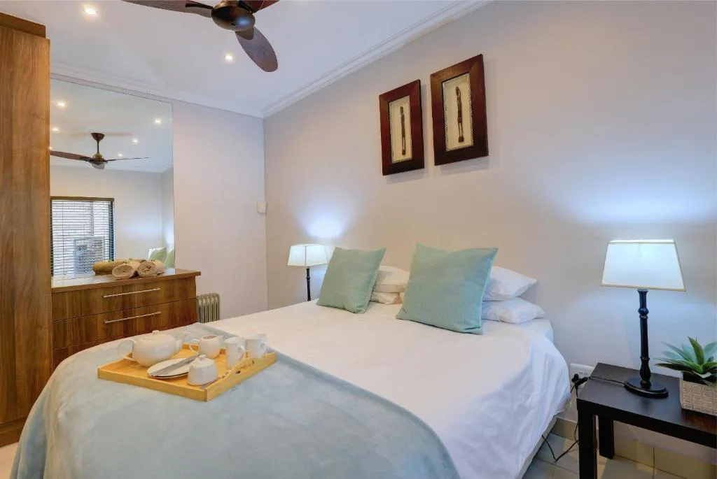 Bright bedroom with white bedding, green cushions, wooden dresser, and side lamps