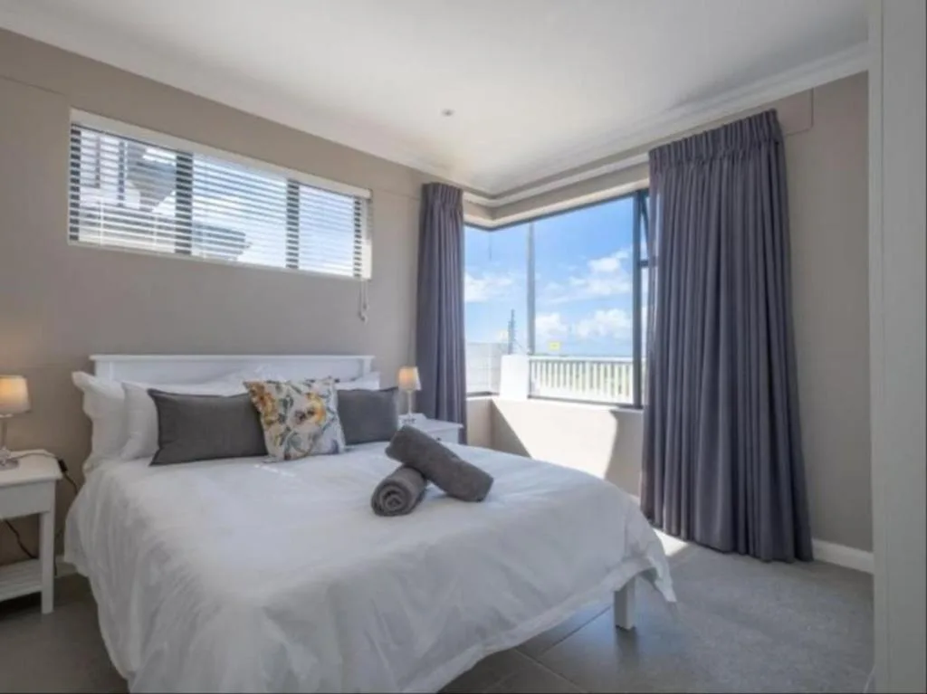 Bright bedroom with white bed, grey pillows, and views of town through large windows