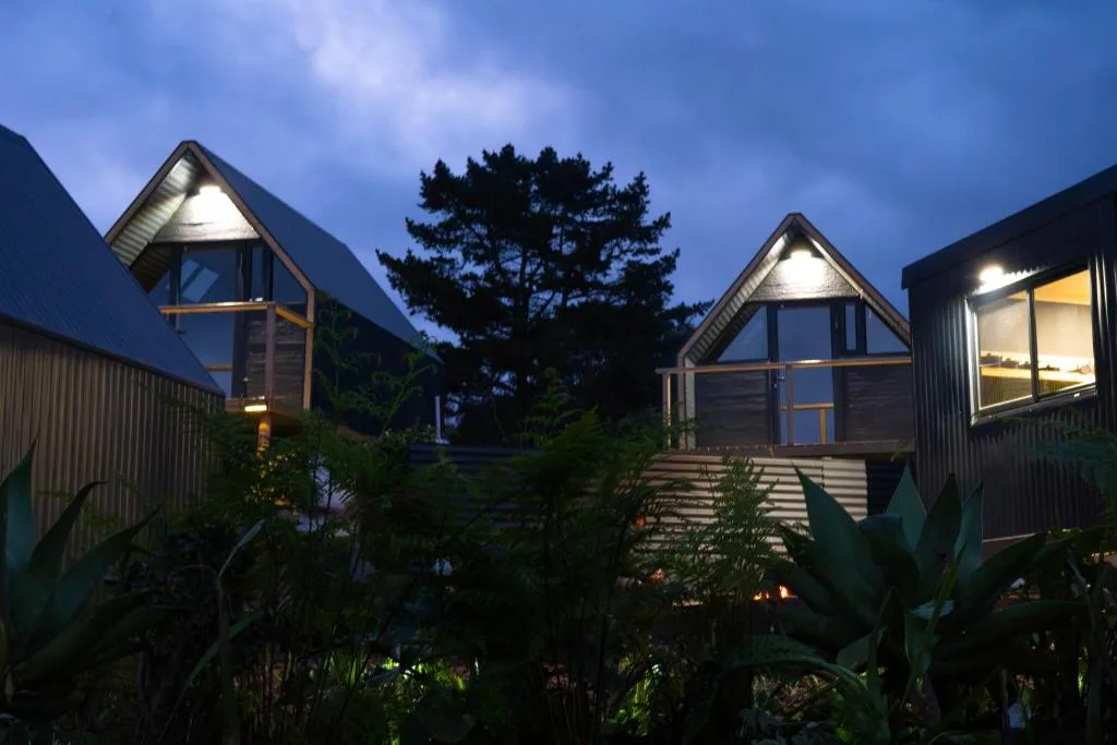 Modern glamping chalets with peaked roofs illuminated at dusk, surrounded by lush garden vegetation