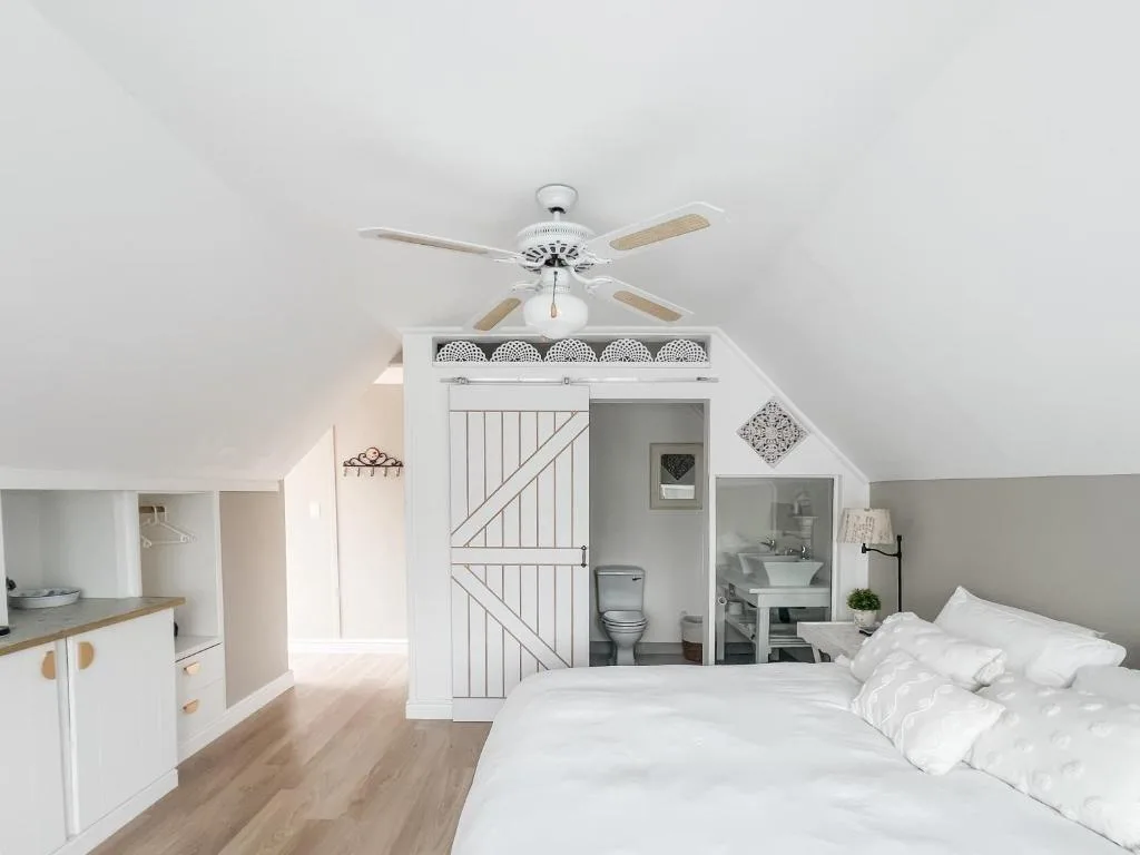 Bright attic bedroom with white bed, ceiling fan, and ensuite bathroom