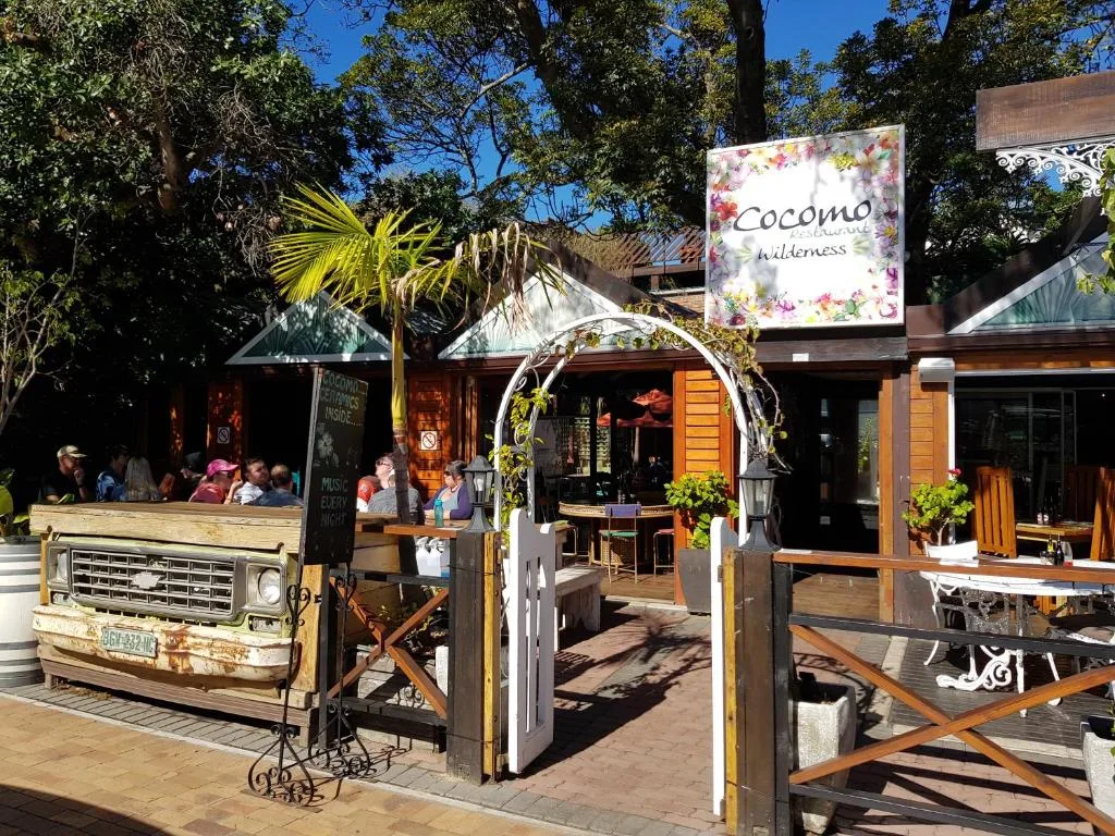 Rustic lodge entrance with signage, outdoor seating, and lush garden surroundings