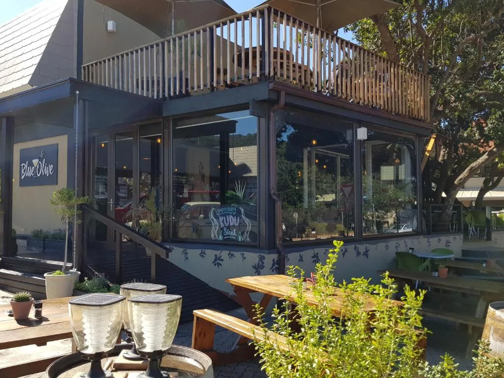 Modern blue cottage with wooden deck, large windows, and green vegetation surroundings