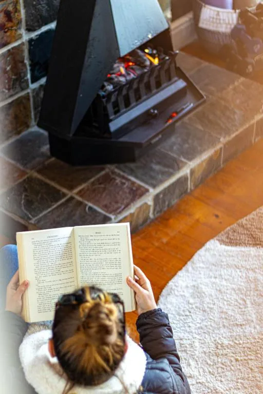 Person reading book by fireplace with glowing flames in cozy interior