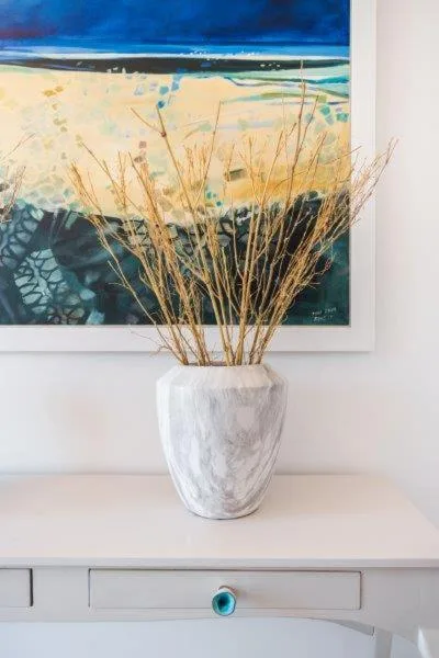 White ceramic vase with dried grasses on white desk beside beach artwork