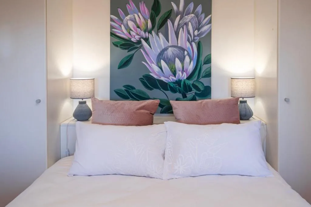 Neatly made bed with white pillows and pink cushions, framed by floral artwork