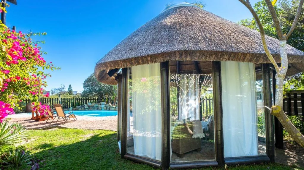 Thatched-roof gazebo with pool and gardens visible beyond bright sunny day
