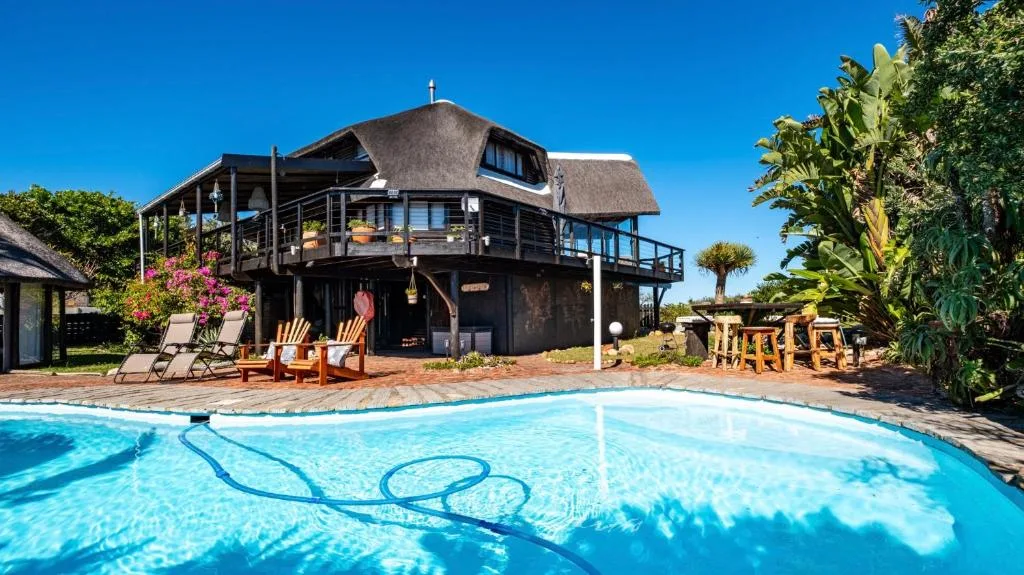 Crystal-clear swimming pool with thatched-roof house and gardens beyond