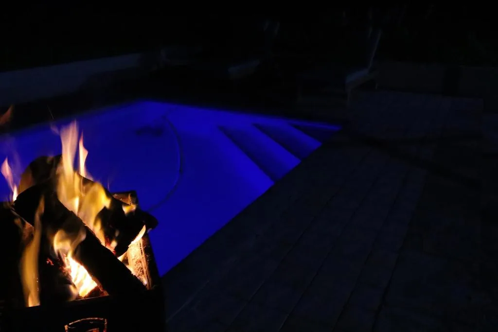 Glowing heated swimming pool lit by blue night lighting beside a fire