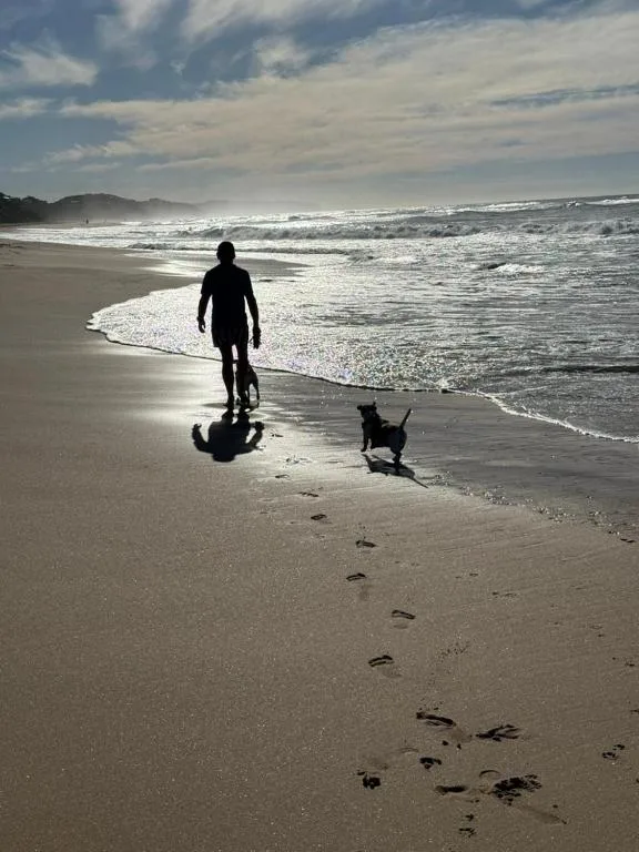 Surfer with dog walking along sandy beach with breaking ocean waves